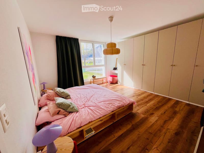 A bedroom with a bed, pillows, and a lamp. There is a nightstand with a lampshade, a painting on the wall, and a curtain on the window. A wardrobe is placed against the wall, and a desk with a chair is near the window. A storage box and a plant are on the