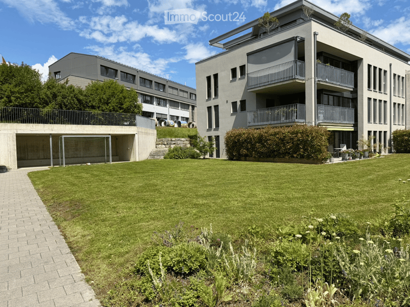 Modern multi-story building with balconies, surrounded by a well-maintained lawn and garden area. Includes a covered garage and raised ground floor.