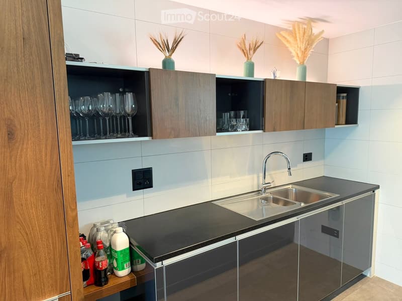 Modern kitchen with light blue and white tiled walls, dark countertops, stainless steel sink, and wooden cabinets. Glassware and decorative vases are visible on the shelves.