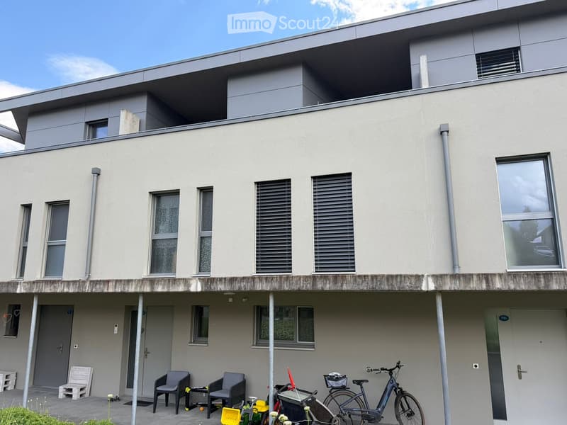 Modern two-story building with a flat roof, large windows, and a raised ground floor. The entrance features a small seating area with chairs and a table, bicycles, and a stroller.