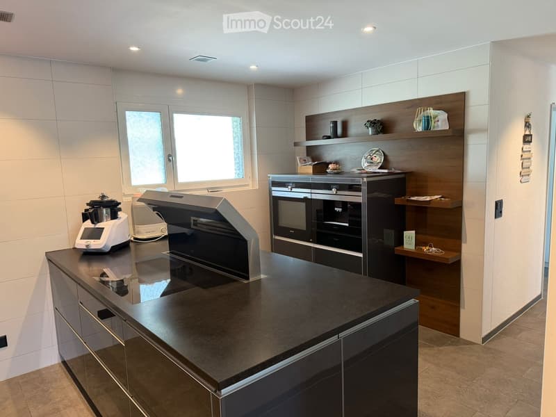 Modern kitchen with dark countertops, built-in appliances, and wooden shelving. Features a blender, oven, and decorative items on shelves.