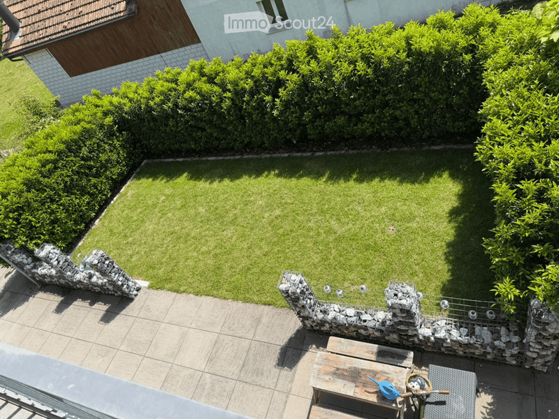 Well-maintained garden with a neatly trimmed lawn, surrounded by hedges and a stone wall. A small wooden bench and gardening tools are visible on the patio area.