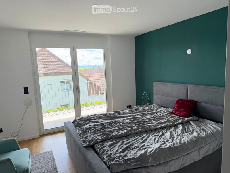Modern bedroom with a large bed, grey headboard, red pillow, grey duvet, a teal armchair, and a table lamp on a nightstand. Large windows offer a scenic view of the surrounding area.