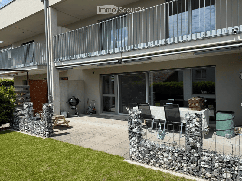 Outdoor patio area with a table, chairs, grill, and stone walls, adjacent to a grassy lawn and a modern building with balconies.