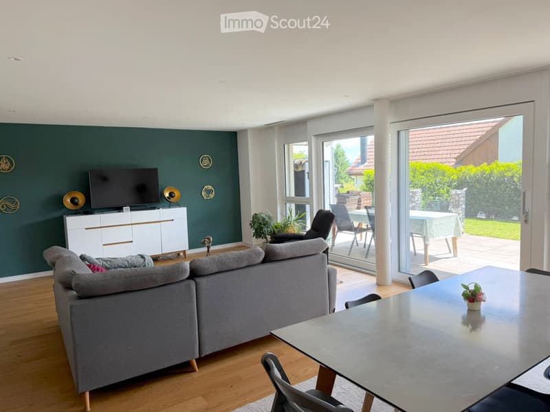 Modern living room with grey sectional sofa, glass coffee table, TV on white cabinet, and large sliding glass doors leading to a patio with outdoor furniture.
