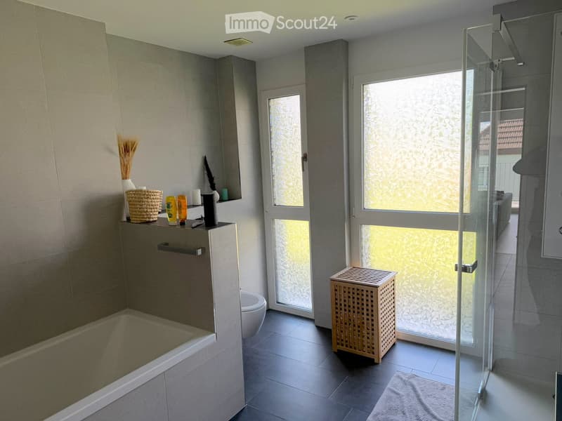 Modern bathroom with a bathtub, toilet, glass-enclosed shower, and decorative items including a wicker basket, candles, and a wooden stool.