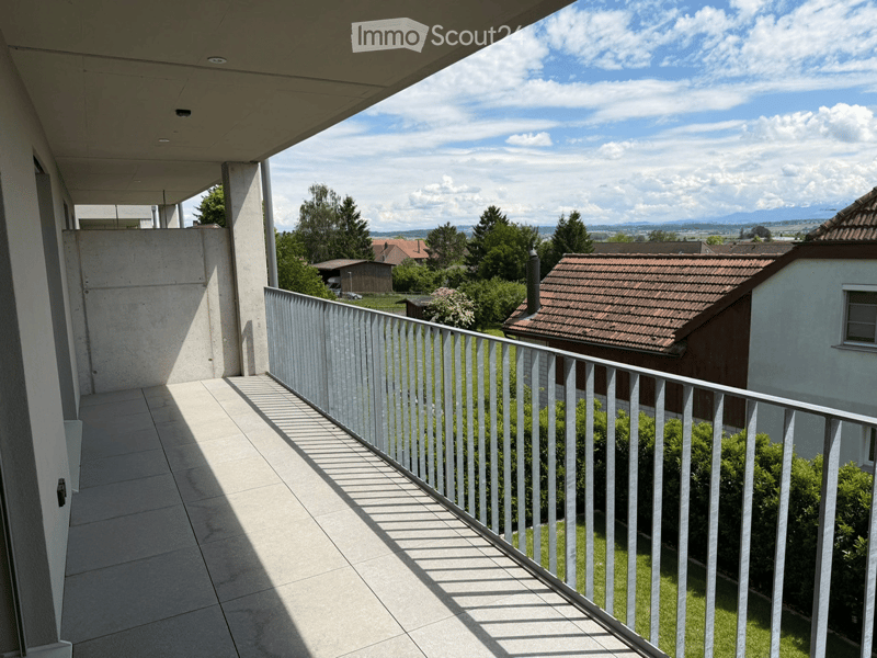 Balcony with tiled floor, white metal railing, overlooking a green landscape with trees and houses.