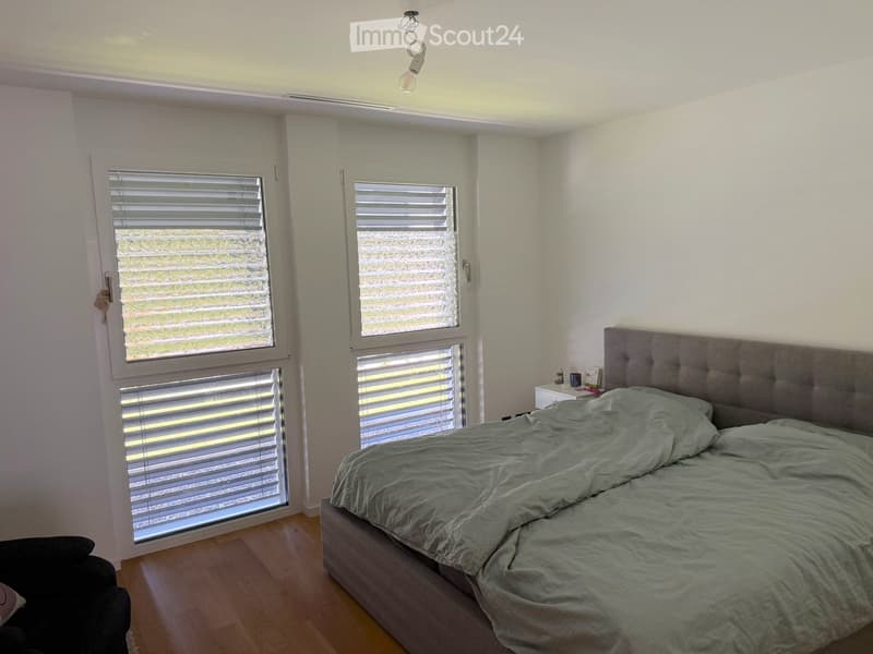 Bright bedroom with wooden flooring, large windows with blinds, a grey upholstered bed, and a nightstand with a lamp.