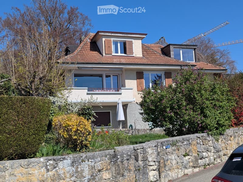Two-story house with a tiled roof, multiple windows, and a balcony on the first floor, surrounded by greenery and a stone wall.