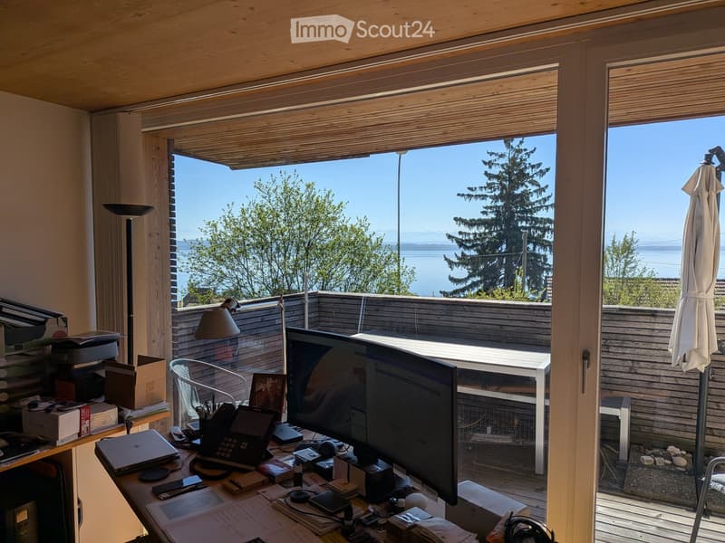 A modern, well-lit room with a large window offering a scenic view of the water and trees. The room features a desk with a laptop, a screen, and various office supplies, along with a chair and a lamp. An umbrella is visible near the window, and a small ta