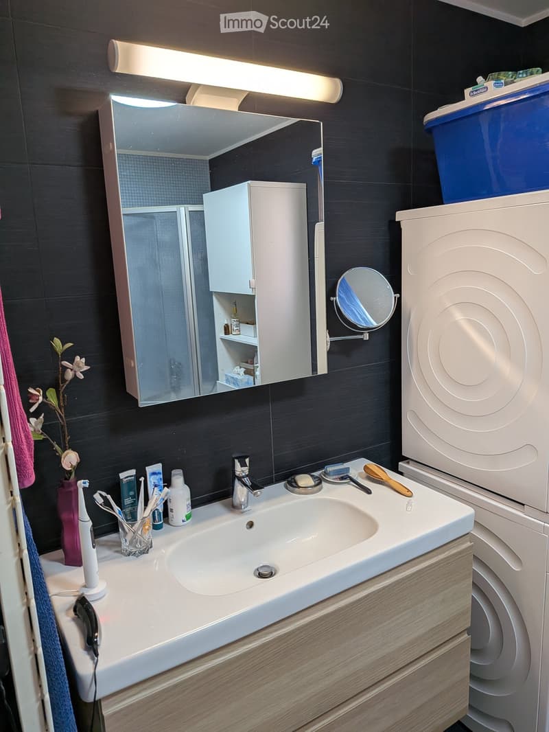 Modern bathroom with a white sink, mirror, and wooden cabinet. The sink has a modern faucet and is surrounded by various toiletries including a toothbrush holder, toothpaste, and an electric toothbrush. A towel hangs on the left side, and a small flower v