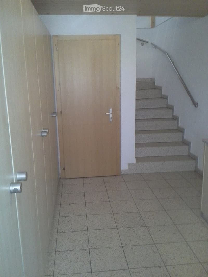 Empty hallway with tiled floor, wooden door, and staircase leading upwards with a metal railing.