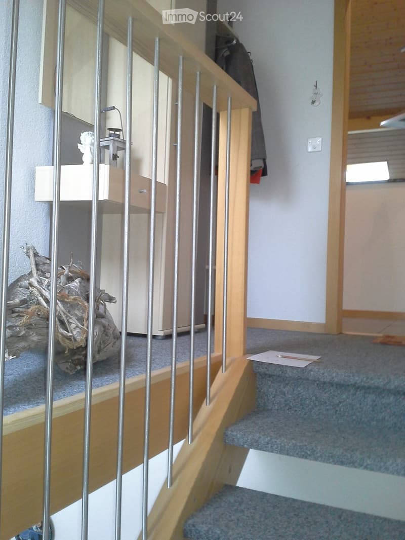 Staircase with wooden handrail and metal balusters, carpeted steps, small shelf with decorative items, coat hanging on wall, and a piece of paper on the floor.