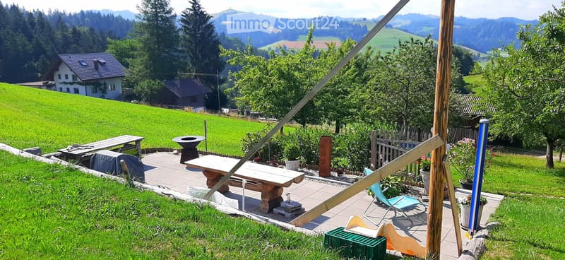 A serene garden area with a tiled patio, wooden bench, table, chairs, and flower pots, surrounded by lush greenery and a scenic mountain view.