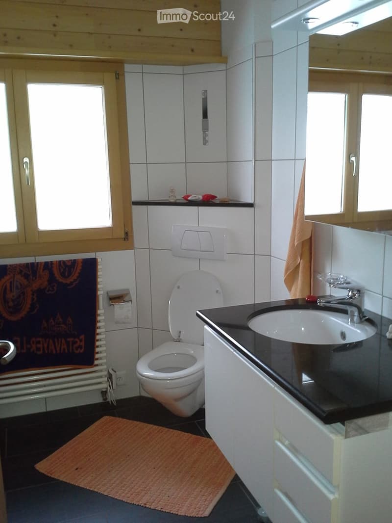 Bathroom with white tiled walls, a toilet, a sink with a mirror, a towel, and a small shelf above the toilet.