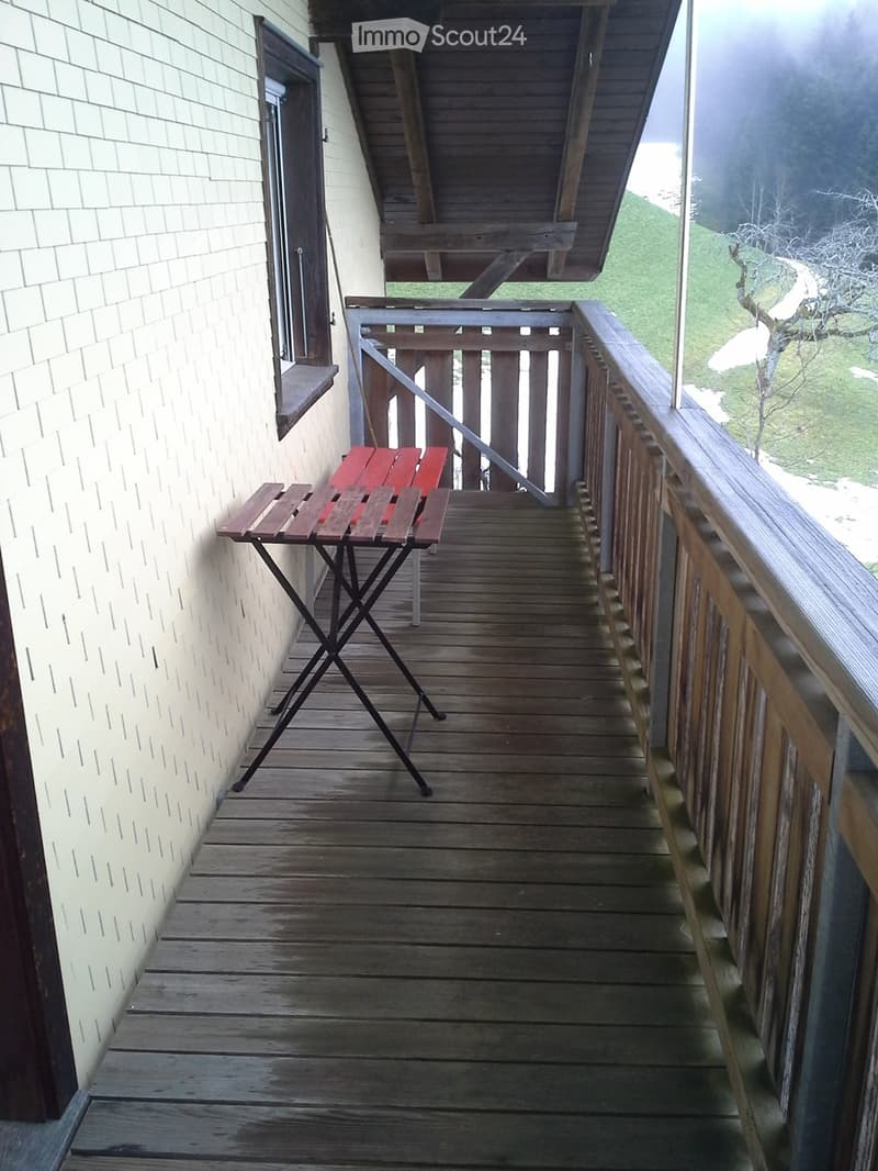 Wooden balcony with a small red table and chair, overlooking a grassy area with trees.