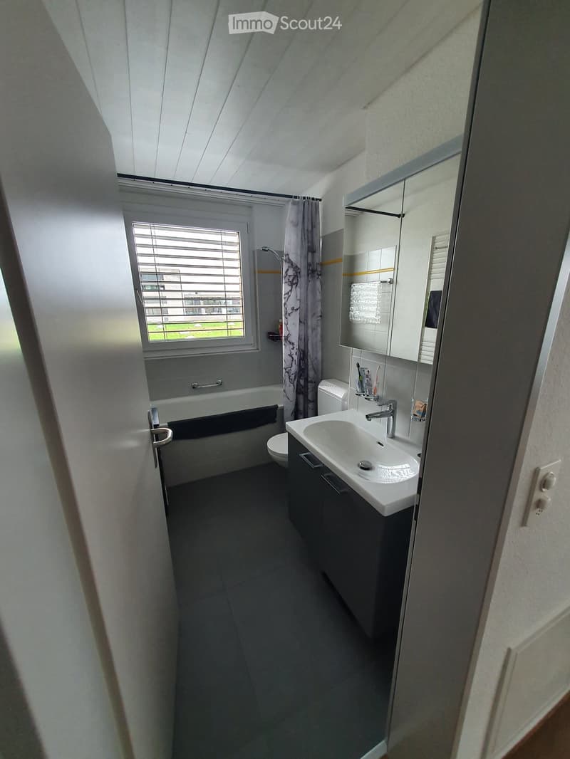 Bathroom with a bathtub, sink, toilet, and shower area. The sink has a faucet and a mirror above it. The shower area is covered with a curtain, and there is a shower head attached to the wall. The bathroom has a white ceiling and walls, and the floor is t