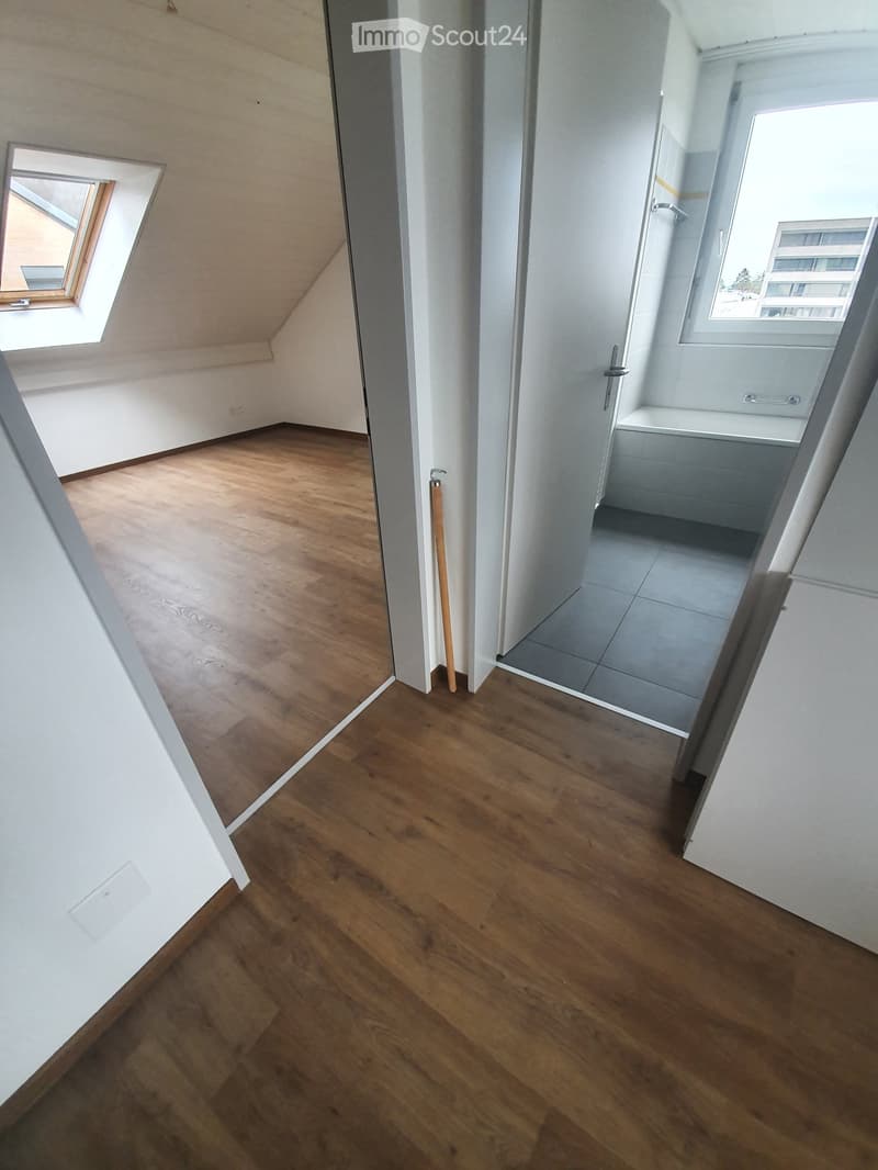 Empty room with wooden flooring, white walls, and a door leading to a bathroom. There is a window on the ceiling.