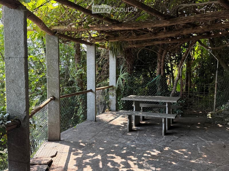 An outdoor patio area with a stone floor, wooden pergola, and a table with benches, surrounded by lush greenery and a chain-link fence.