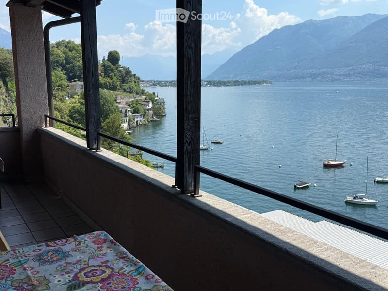Balcony with a scenic view of a lake and mountains, featuring a tiled floor and a colorful tablecloth on a small table.