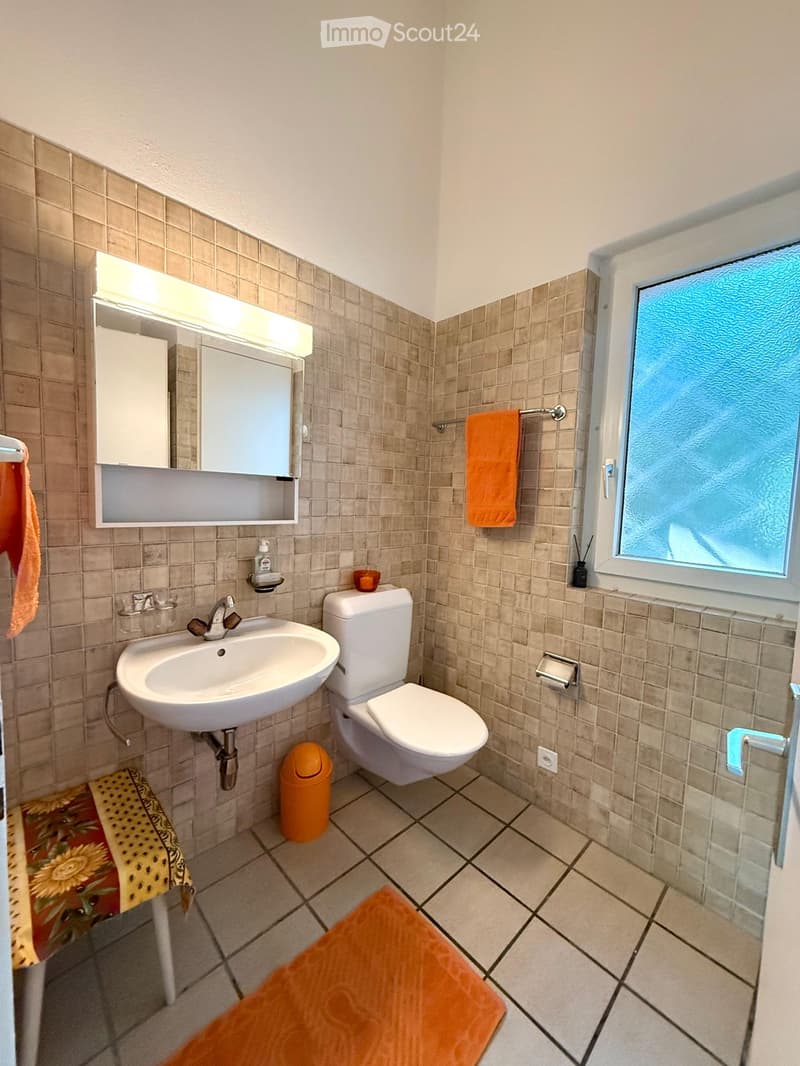 Bathroom with beige tiled walls and floor, white sink, toilet, mirror with built-in cabinet, and orange towels.