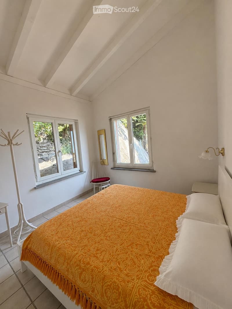 Cozy bedroom with a slanted ceiling, large windows, a bed with an orange bedspread, a small chair, a mirror, and a coat rack.