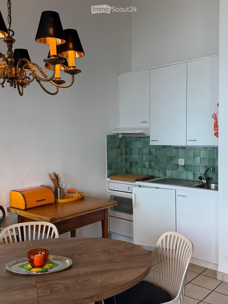 A modern kitchen with white cabinets, a wooden dining table, and a chandelier. The kitchen features a sink, oven, and various appliances.
