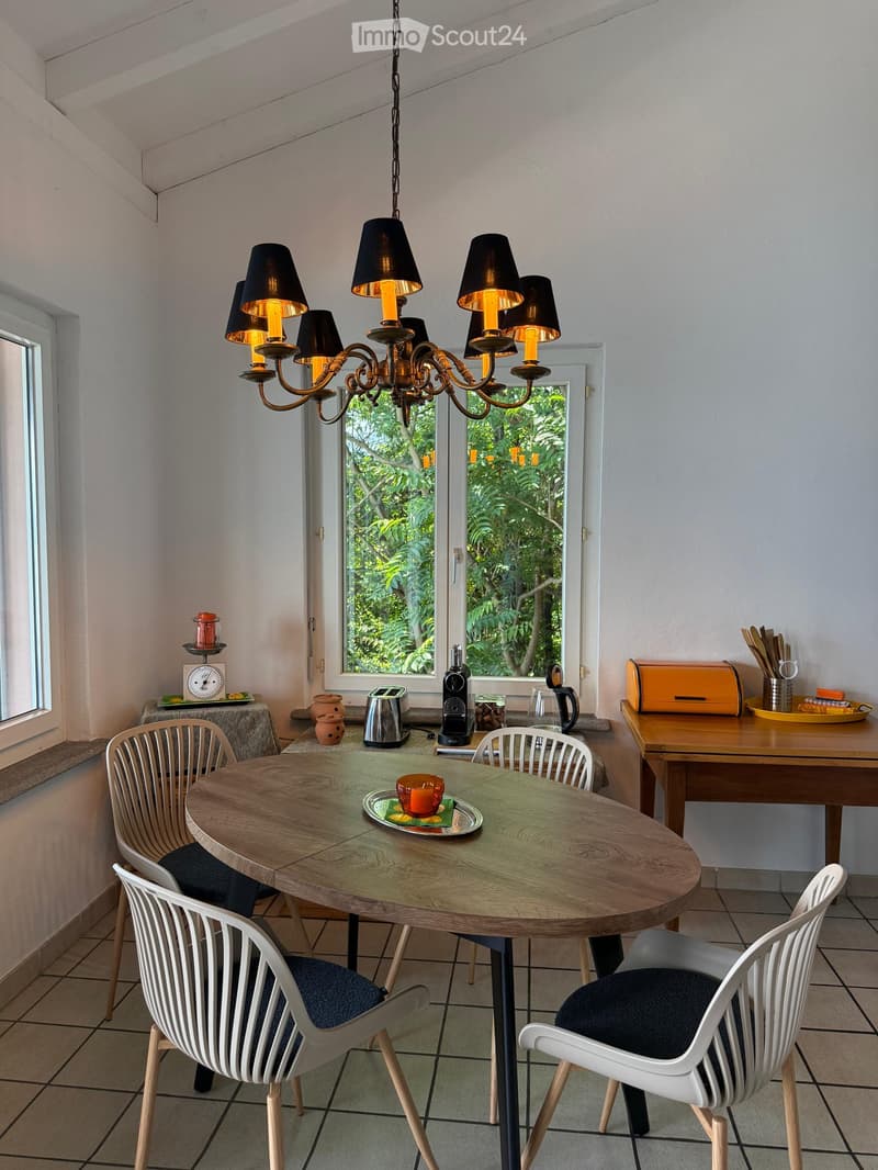 Bright kitchen with a round wooden dining table, four white chairs, a coffee maker, toaster, and a small cabinet with kitchen utensils.