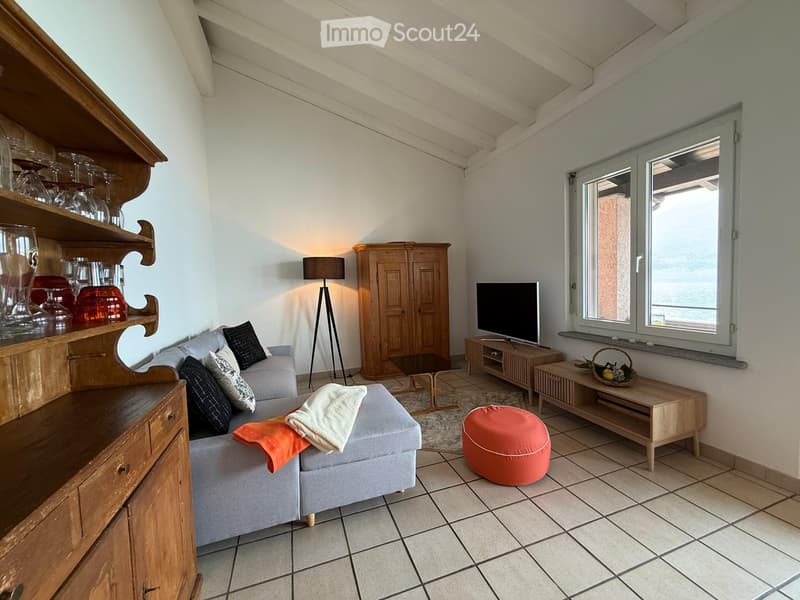 A cozy living room with a grey sofa, ottoman, wooden cabinet, television, floor lamp, and a shelf with glassware. The room features large windows with a scenic view and tiled flooring.