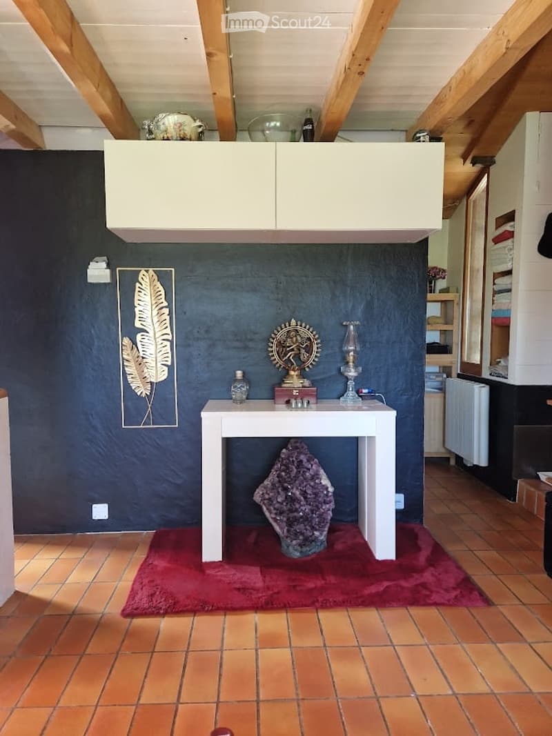 A small, furnished room with a white table, a large amethyst crystal, a decorative statue, a candle holder, and a picture frame on the wall. The room features exposed wooden beams on the ceiling and a dark blue accent wall.