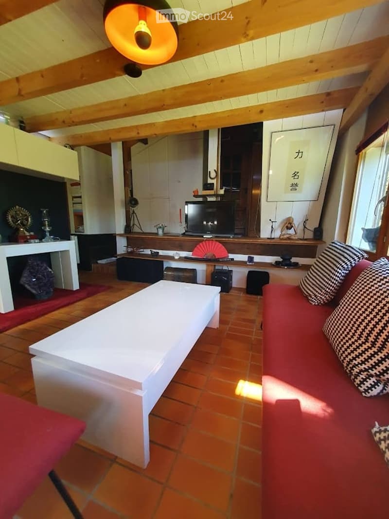 Living room with exposed wooden beams, red sofa, white coffee table, fireplace, and television. The room features a warm, inviting atmosphere with a mix of modern and traditional decor.