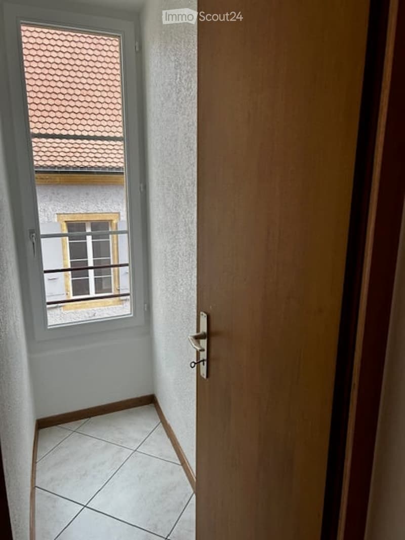 A narrow hallway with tiled flooring, white walls, and a wooden door on the right. A window on the left shows a view of a tiled roof and part of a neighboring building.
