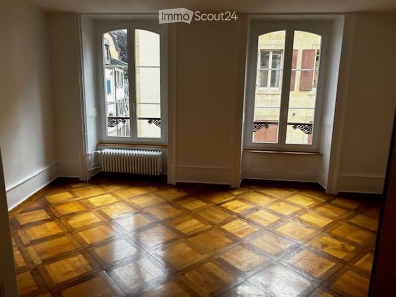 Empty room with wooden parquet flooring, large windows, radiator, and white walls.