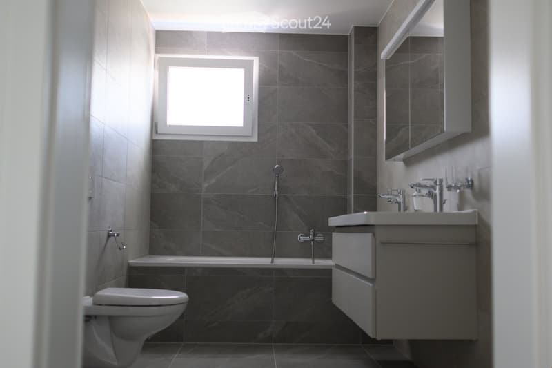 Large bathroom with grey tile walls, a bathtub, a toilet, and a vanity with a sink and mirror.