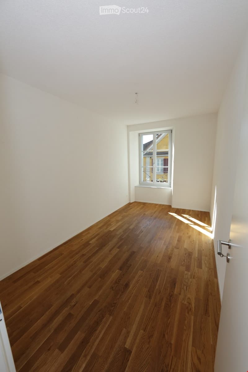 Empty room with light-colored walls and wooden flooring, featuring a single window with a view of a neighboring building.