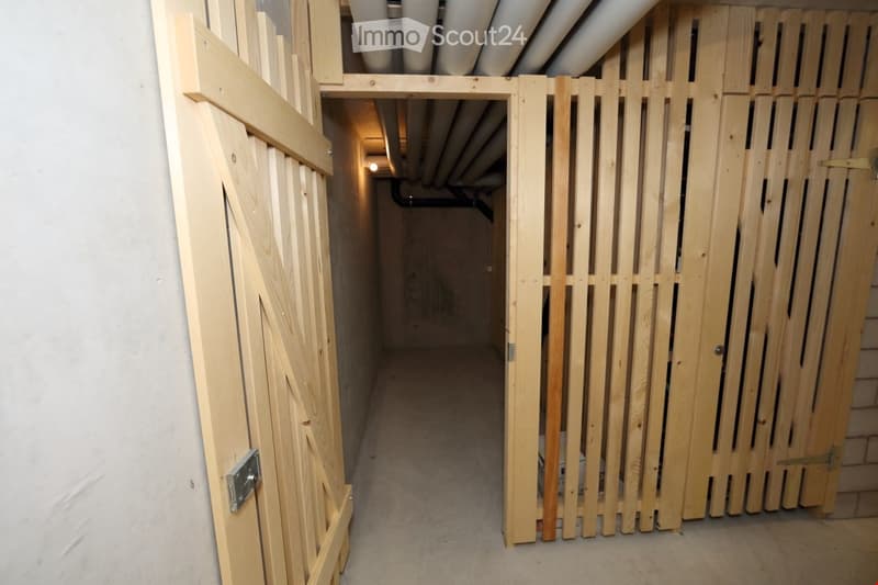 A narrow, unfinished hallway with wooden doors and exposed pipes on the ceiling. The floor is carpeted, and the walls are plain.