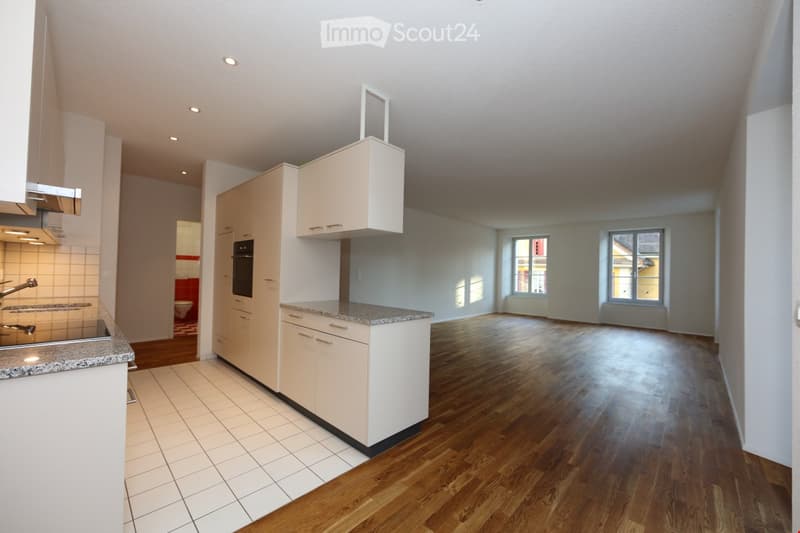 Empty room with wooden flooring, white walls, and large windows. The kitchen area features white cabinets, a granite countertop, and stainless steel appliances.