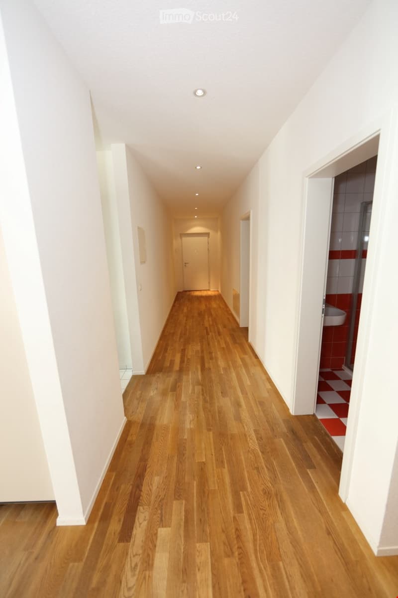 A long, well-lit hallway with wooden flooring, white walls, and recessed ceiling lights. A door is visible at the far end, and a small bathroom is partially visible on the right side.