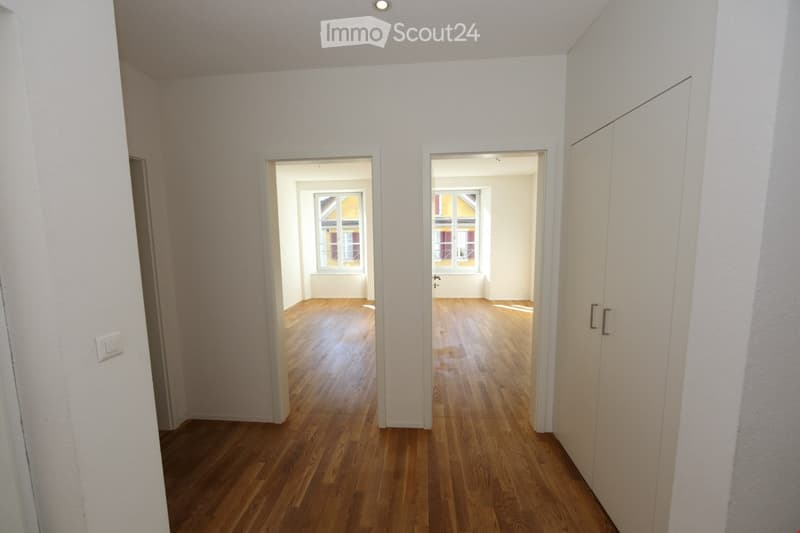 Empty hallway with wooden flooring, white walls, and two open doorways leading to adjacent rooms with windows.