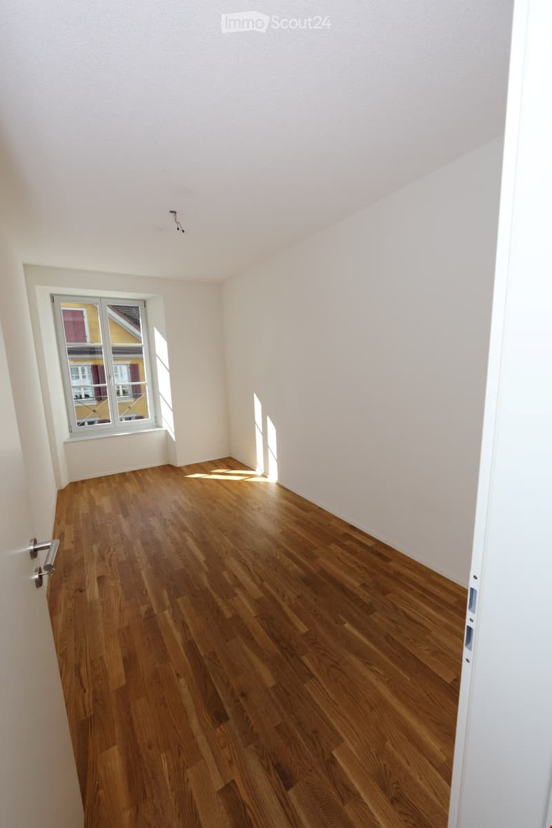 Empty room with wooden flooring and white walls, featuring a single window with a view of neighboring buildings.
