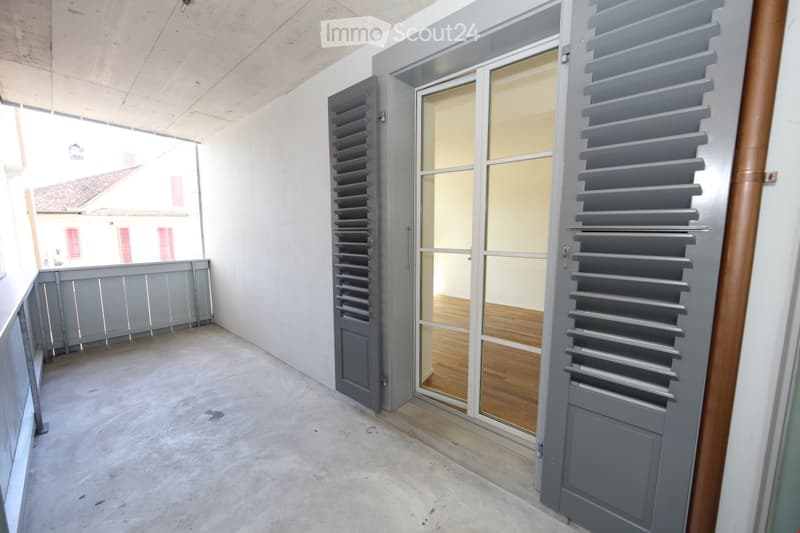 Empty balcony with concrete floor, grey shutters, and a sliding glass door leading to an interior space with wooden flooring.
