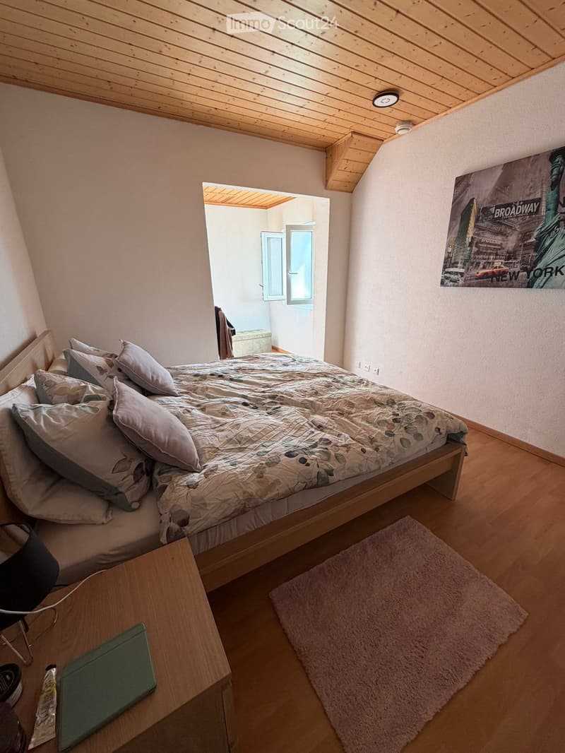 Cozy bedroom with a wooden bed, nightstand, chair, laptop, and a large poster on the wall. The room features wooden flooring and a slanted wooden ceiling.