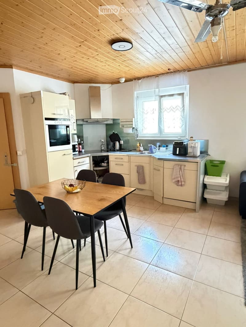 Modern kitchen with light wood cabinets, stainless steel appliances, a dining table with four chairs, and a window with blinds.