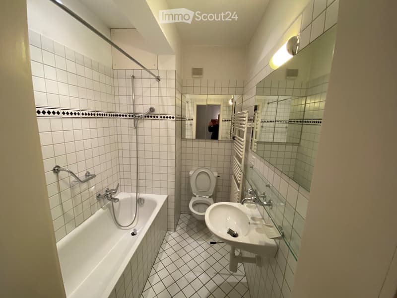 Bathroom with white tiled walls and floor, featuring a bathtub, toilet, sink, mirror, and radiator.