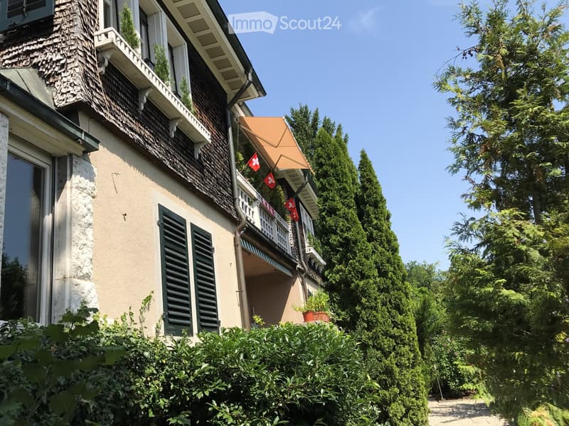 Two-story house with beige walls, dark green shutters, and a shingled roof. Features a balcony with railings and Swiss flags, surrounded by lush greenery and trees.