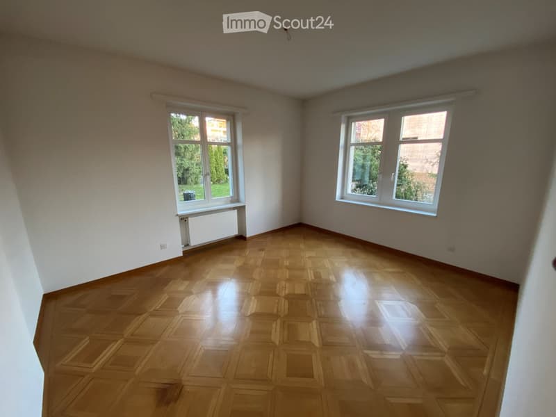 Empty room with wooden parquet flooring, white walls, and large windows offering a view of greenery outside.