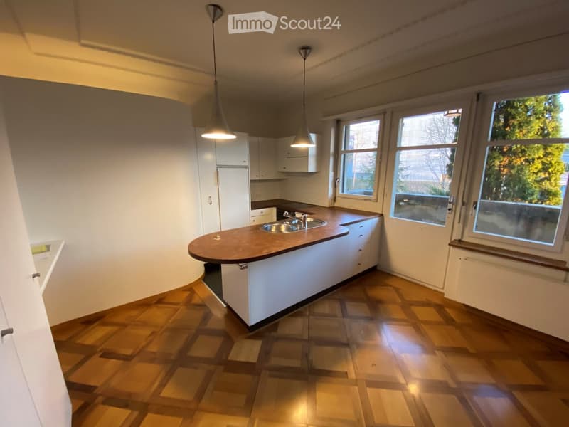 Modern kitchen with white cabinets, a central island, and a polished wooden floor. The island features a countertop and a sink with a faucet. Pendant lights hang above the island.