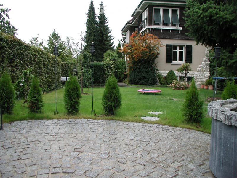 Well-maintained garden with a cobblestone pathway, trimmed hedges, a small trampoline, and a two-story house with multiple windows and shutters in the background.
