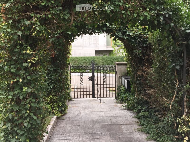 A pathway leads to a gated entrance surrounded by lush greenery and climbing vines, with a modern building visible in the background.