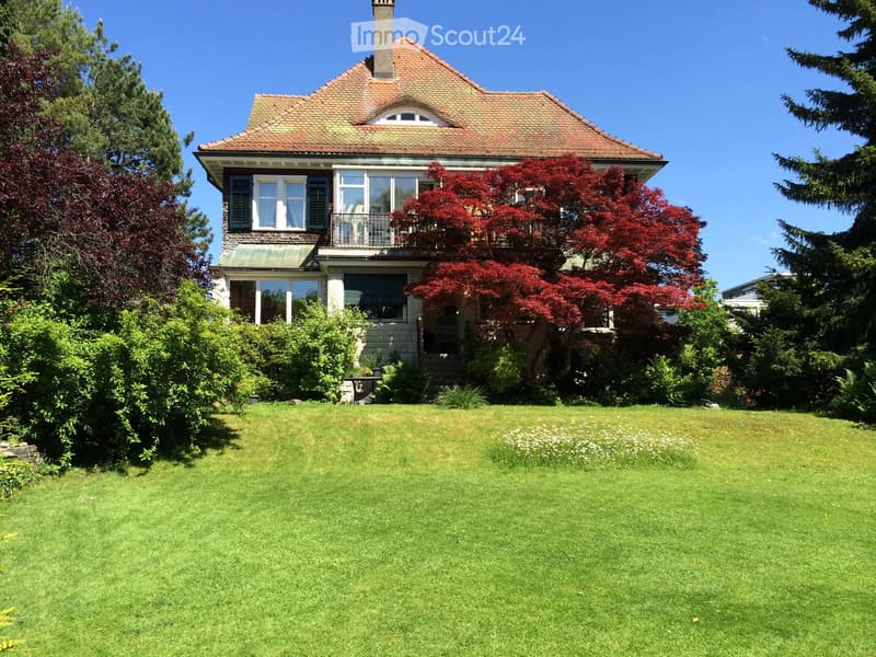 Two-story house with red-tiled roof, large windows, and a well-maintained lawn with trees and shrubs.